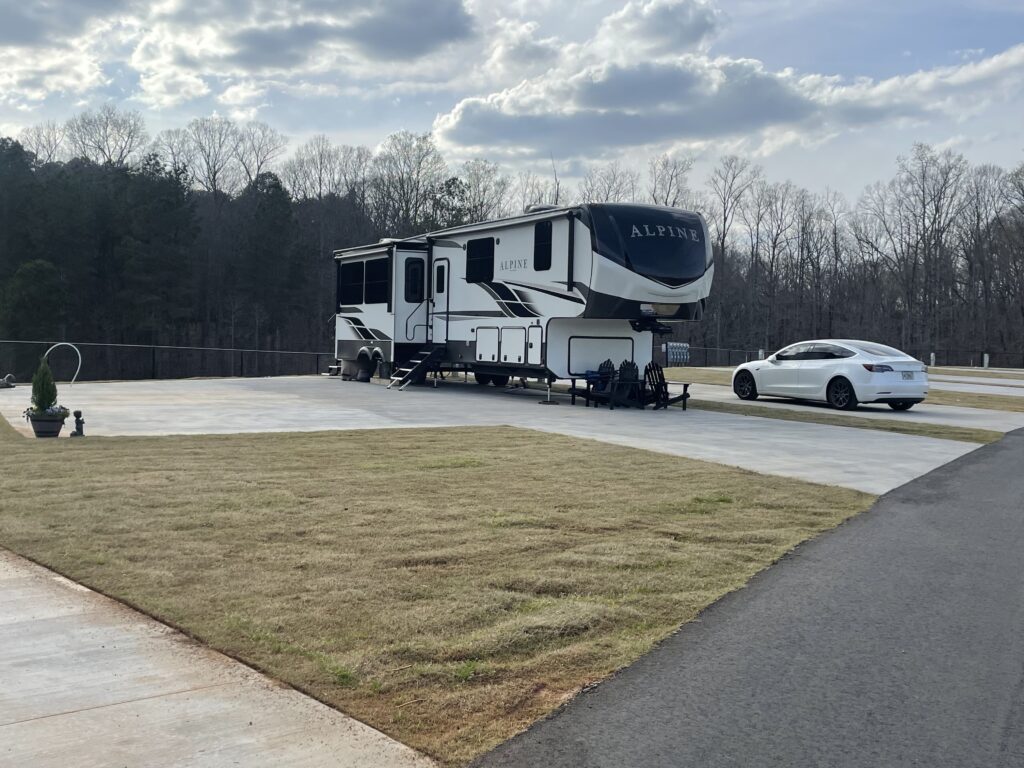 Alpine fifth-wheel RV parked on a concrete RV pad with an adjacent white car at Clemson RV Park, with grass lawn, asphalt road, and wooded background.