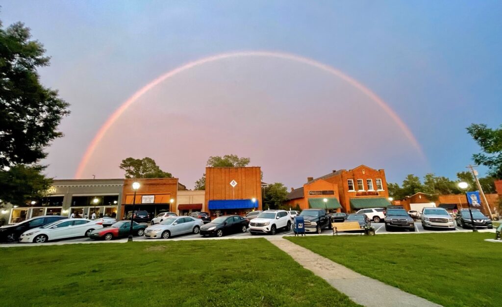 Rainbow over a historic small-town square with a green lawn and brick storefronts